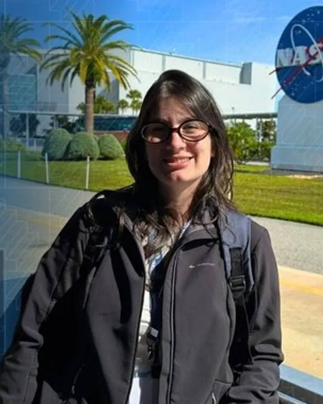 Camila, da Desenrola, sorrindo diante do Kennedy Space Center, com o logotipo da NASA e palmeiras ao fundo