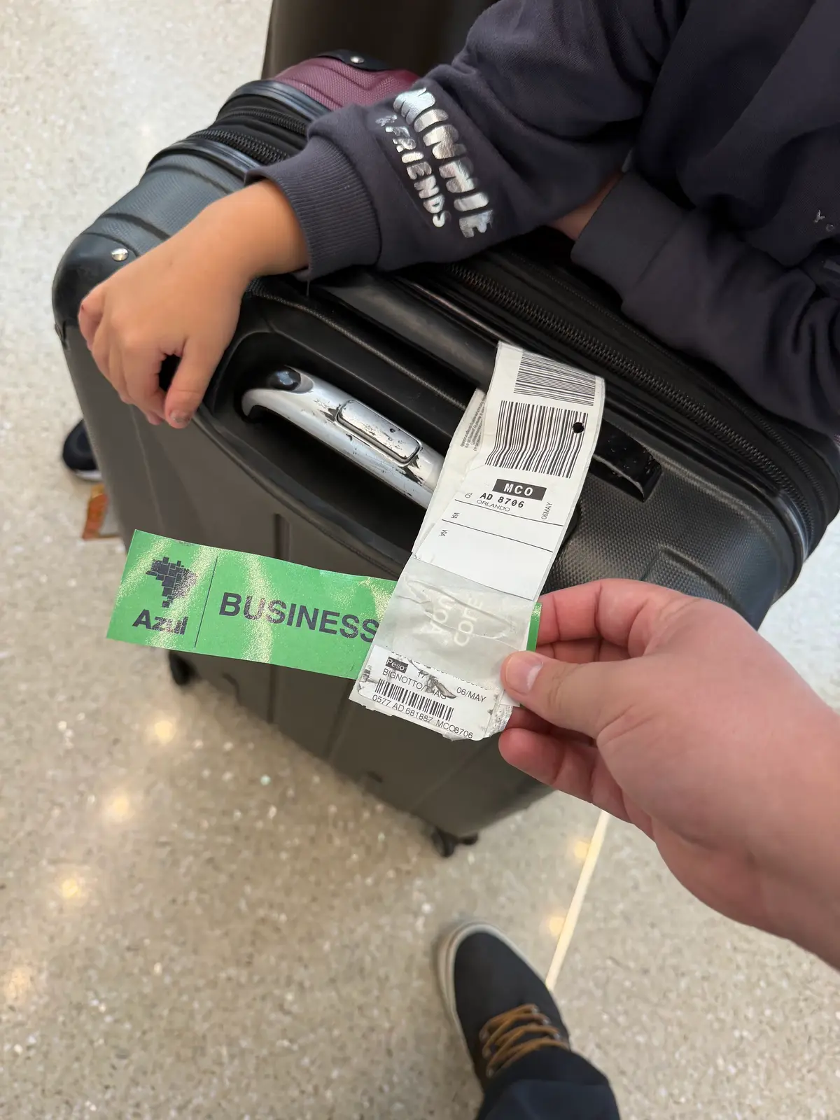 Mão segura a etiqueta de bagagem com destino a Orlando (MCO) ao lado de uma mala de rodinhas, no aeroporto