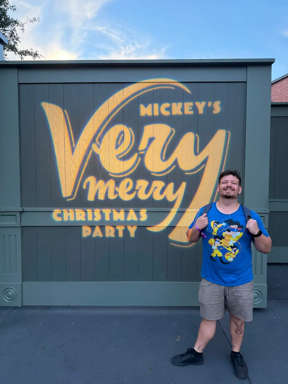 Thomaz com camiseta azul do Pateta fazendo joinha em frente ao painel da Mickey's Very Merry Christmas Party, no Magic Kingdom